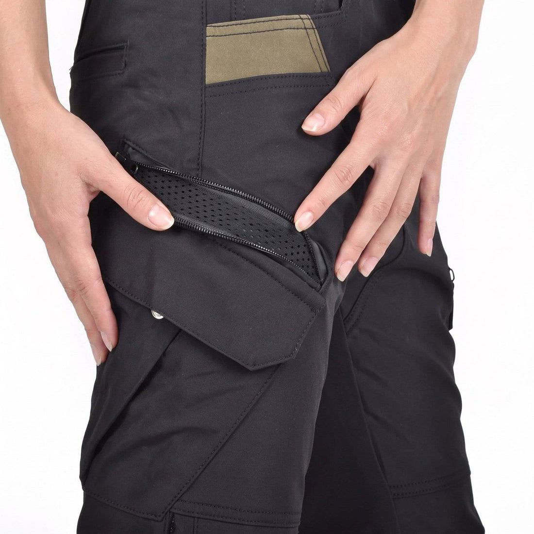Zwei Hände öffnen den schrägen Belüftungsreißverschluss über der Cargotasche einer schwarzen Hose; darunter liegt gelochtes schwarzes Mesh, oben sitzt ein olivfarbener Einsatz