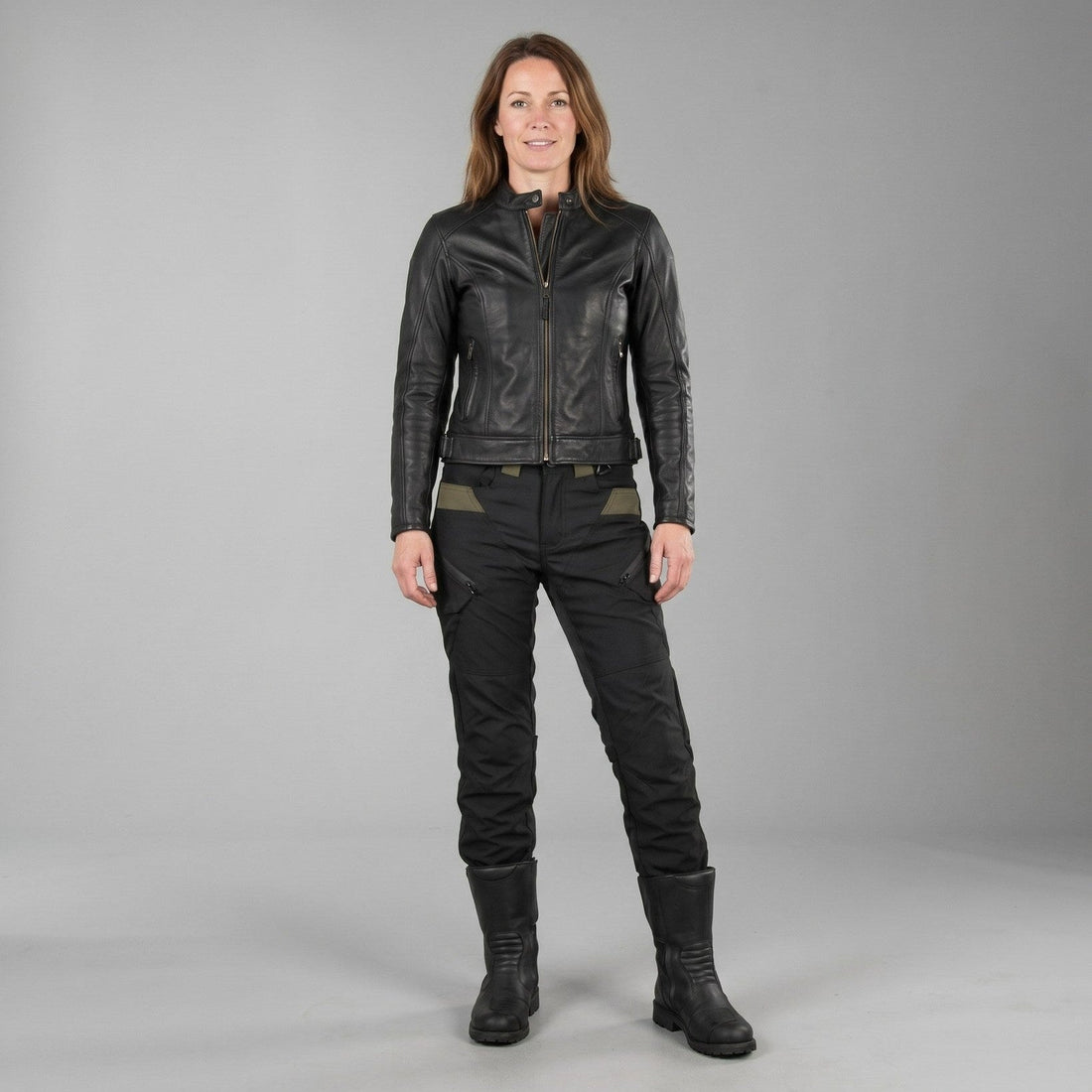 Frau in schwarzer Lederjacke, schwarzer Motorradhose mit oliven Einsätzen und hohen schwarzen Stiefeln, frontal vor grauem Studiohintergrund
