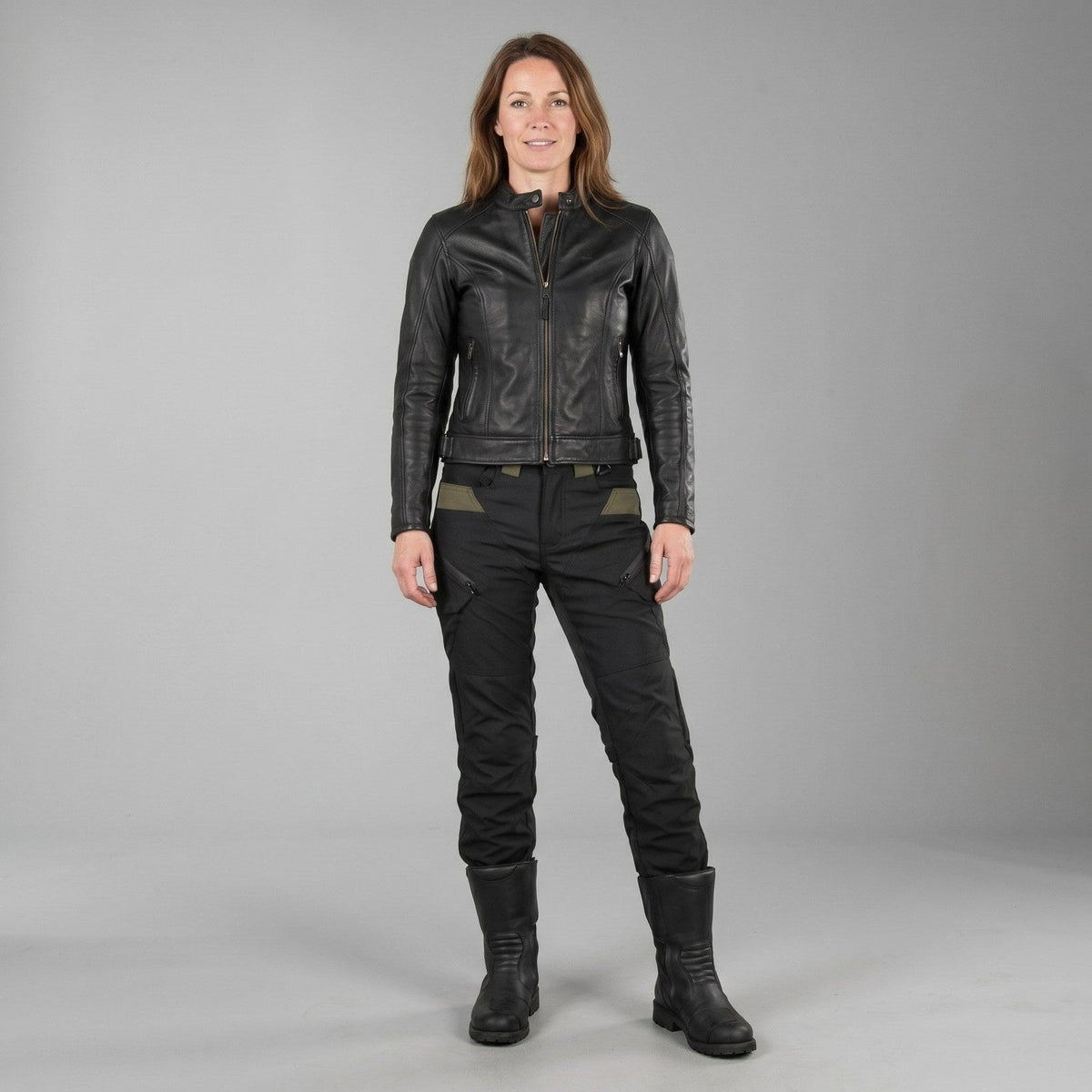 Frau in schwarzer Lederjacke, schwarzer Motorradhose mit oliven Einsätzen und hohen schwarzen Stiefeln, frontal vor grauem Studiohintergrund