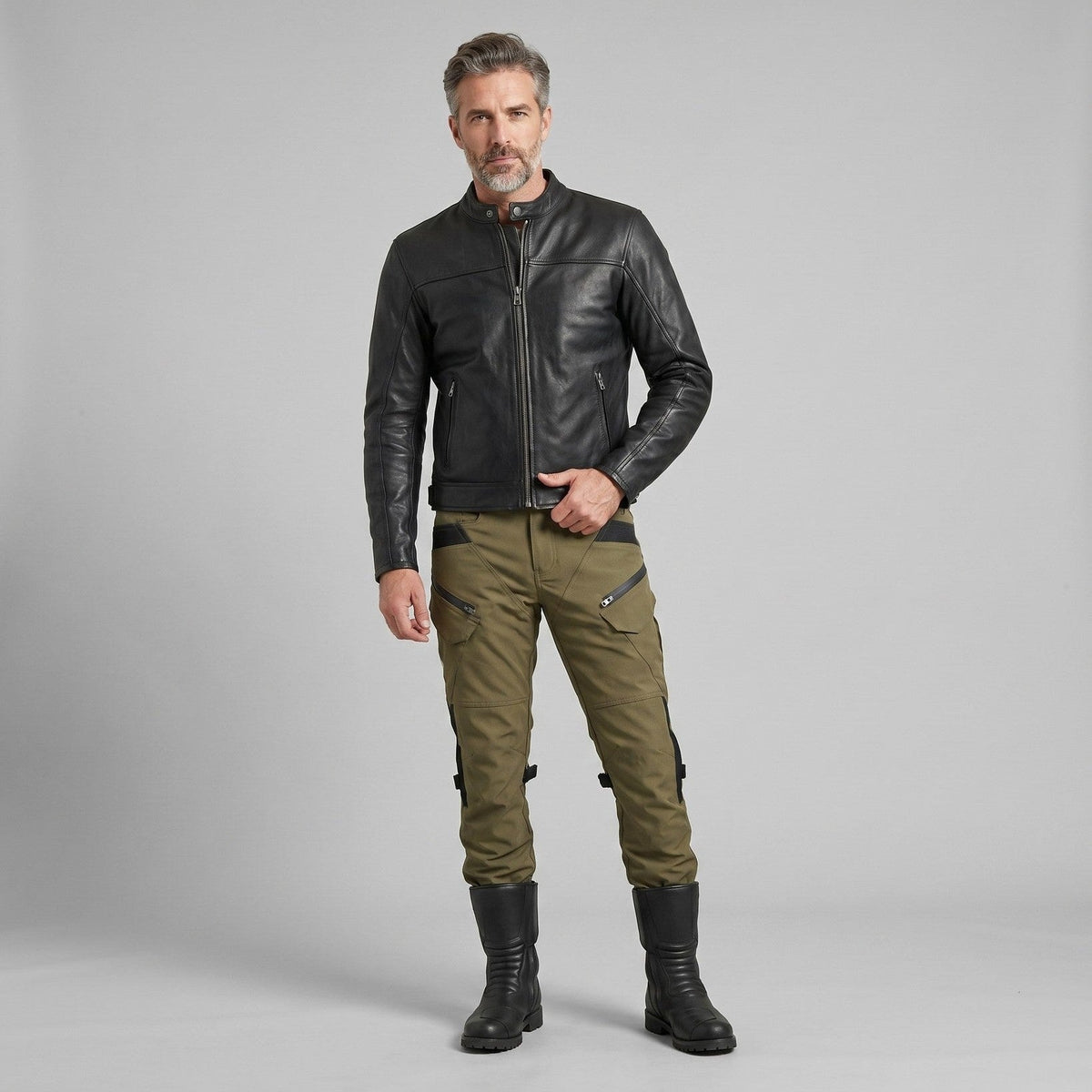 Mann in schwarzer Lederjacke und olivgrüner RoadVault-Motorradhose mit schwarzen Einsätzen, Kniegurten und hohen schwarzen Stiefeln vor grauem Studiohintergrund