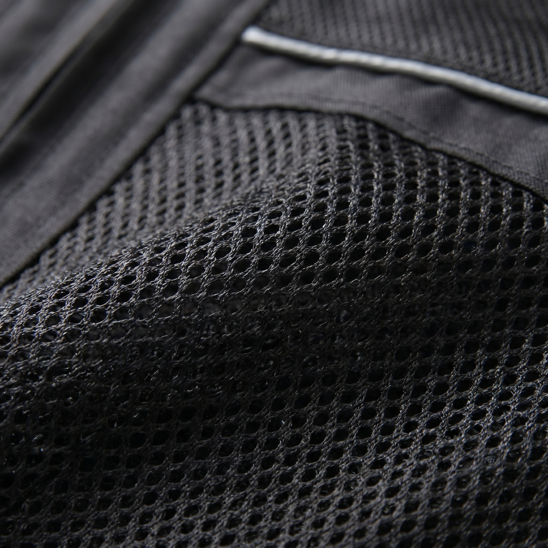 Nahaufnahme des Netzgewebes einer schwarzen Motorradjacke mit silbergrauem Reflexstreifen am oberen Rand und glattem Textil links daneben