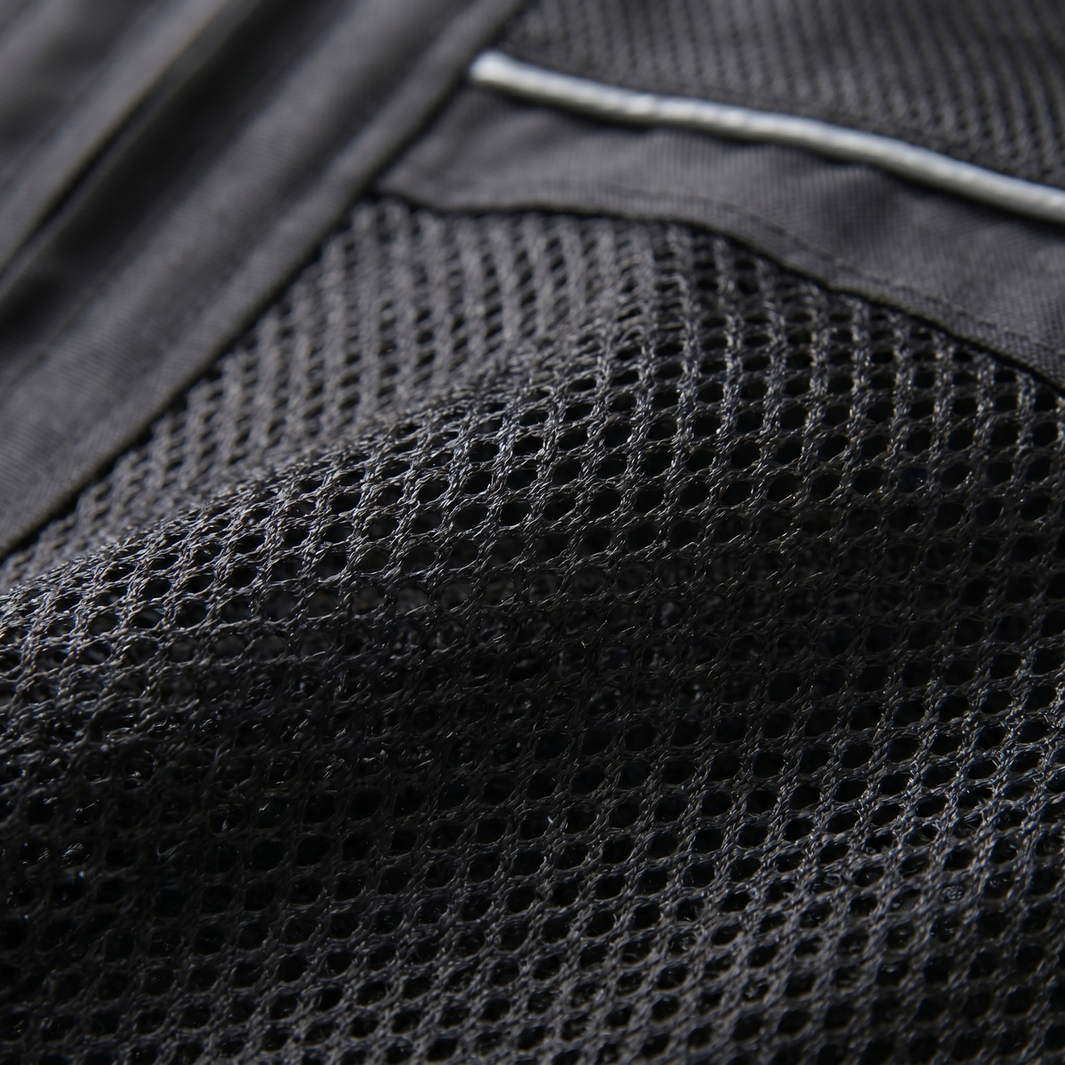 Nahaufnahme des Netzgewebes einer schwarzen Motorradjacke mit silbergrauem Reflexstreifen am oberen Rand und glattem Textil links daneben