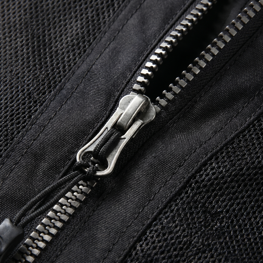 Nahaufnahme des metallenen Reißverschlusses einer schwarzen Mesh-Jacke mit Schieber, geflochtenem Zugband und grobmaschigem Netzgewebe links und rechts der Zahnreihe