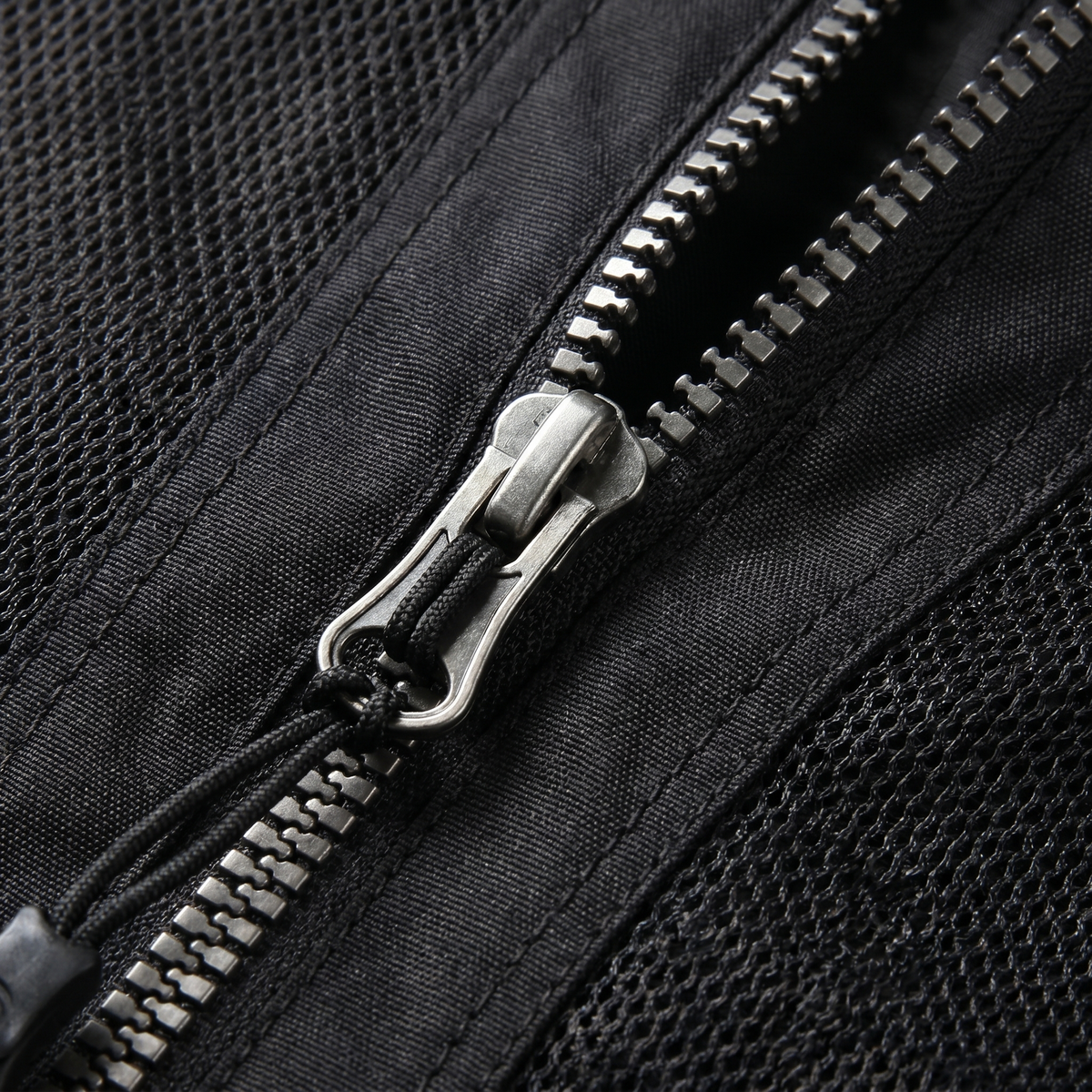 Nahaufnahme des metallenen Reißverschlusses einer schwarzen Mesh-Jacke mit Schieber, geflochtenem Zugband und grobmaschigem Netzgewebe links und rechts der Zahnreihe
